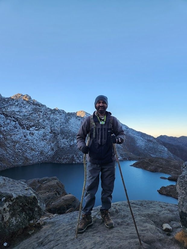 Ajay standing on rocks above a still alpine lake at dawn.