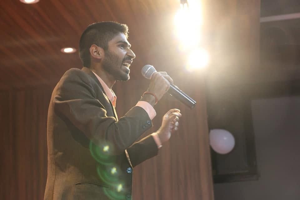 Ajay on stage under warm lights holding a microphone.