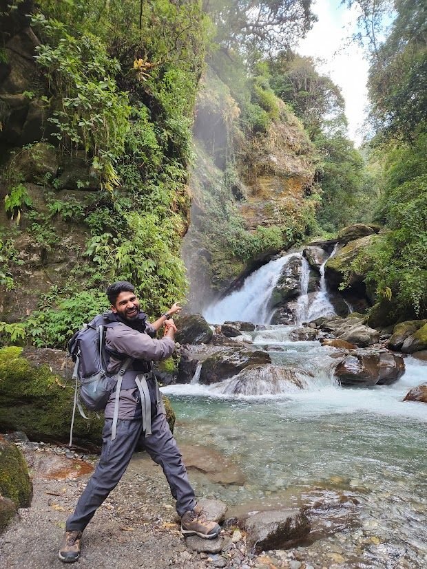 Ajay on a green forest trail beside a waterfall, pack on his back.