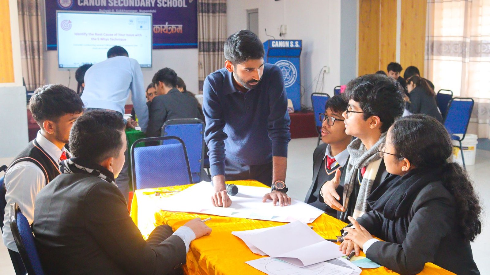 Students in school uniform working around a table while Ajay leans in to listen.