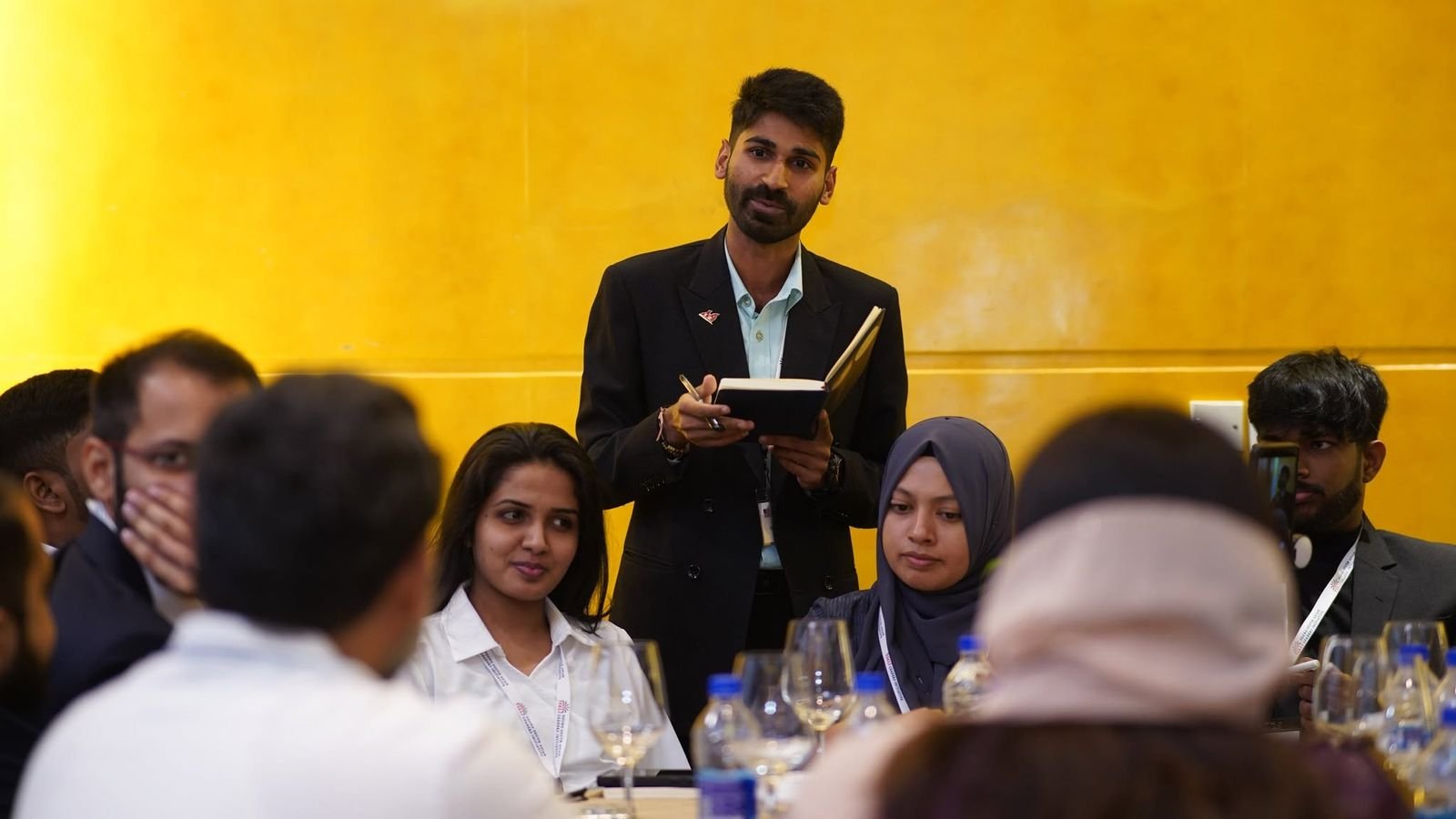 Ajay speaking to a seated group around a table in a bright yellow room.